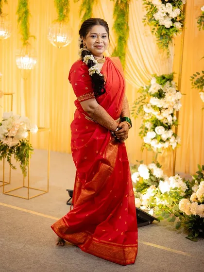A full-length view of a bride in a simple yet elegant red saree, with her hair styled in a traditional floral braid.