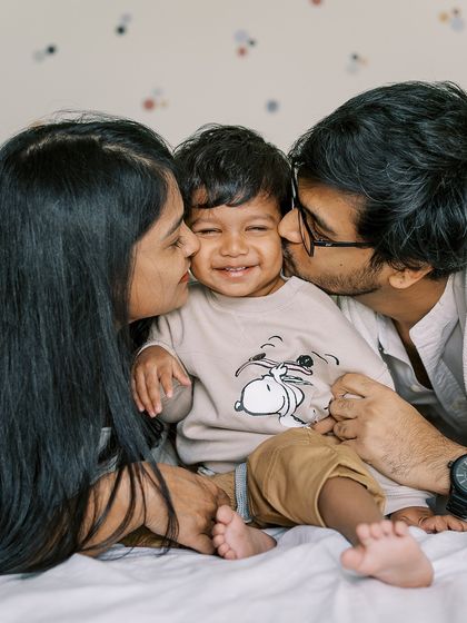 A family sharing a "sandwich kiss" with their smiling son on the bed. These playful moments create the best memories.