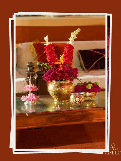 A close-up of a tabletop detail, featuring a traditional brass vase with red flowers and garlands, reflecting the event's regal theme.
