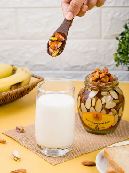 A lifestyle shot showing honey with nuts being added to a glass of milk. This demonstrates a practical and healthy use case for the product.