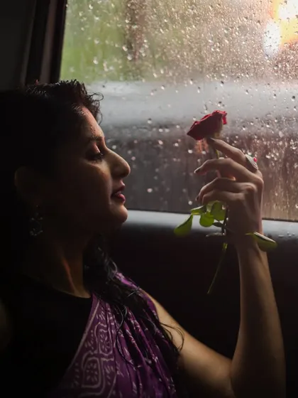 A creative shot from inside a car on a rainy day. The silhouette of the model holding a rose against the raindrop-covered window creates a moody, cinematic, and highly artistic portrait.