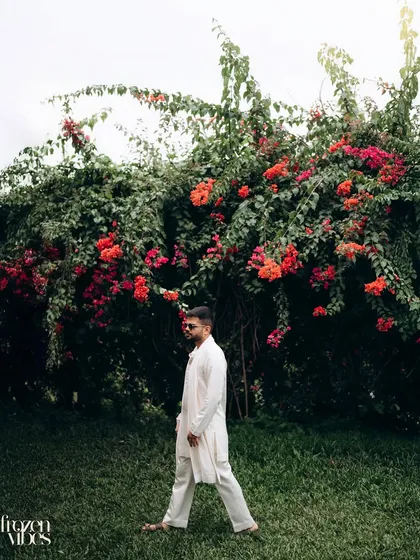 A stylish portrait of the groom, Parth, walking through the lawns of Rhythm Lonavala. The vibrant red flowers of the bougainvillea bush create a stunning natural backdrop.