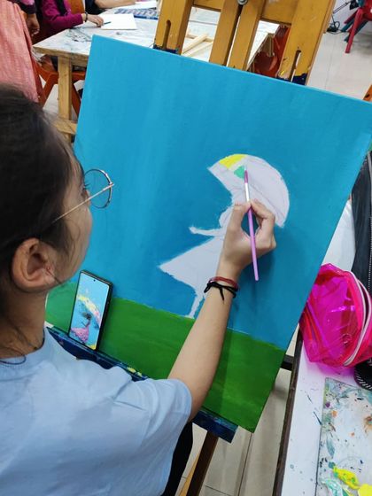 A student works on an imaginative painting of a girl with an umbrella in a field, using a palette knife and brush to create texture.
