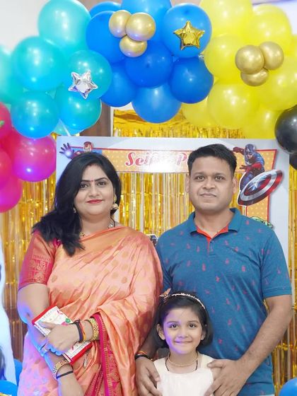 A family of three poses in front of a superhero-themed birthday backdrop with colorful balloons.