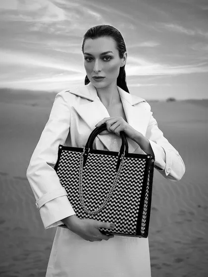 A black and white campaign shot for Da Milano, creating a timeless and classic feel. The model, styled in a white trench coat, holds a geometrically patterned tote bag, with the dramatic sky adding to the cinematic mood.