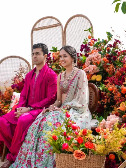 A happy couple at their sun-kissed Haldi in Udaipur. They are seated amidst a stunning explosion of citrus-hued florals, including oranges, pinks, and reds, creating a vibrant and joyful backdrop.