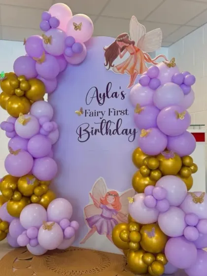 Ayla's "Fairy First Birthday" celebration. This setup features a lovely lavender and gold balloon arch, fairy cutouts, and a personalized backdrop for an enchanting party.