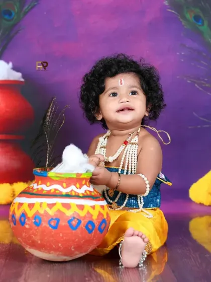 A playful baby with curly hair dressed as Little Krishna, interacting with a pot of "butter." The genuine smile and detailed setup make this a heartwarming and authentic portrait.