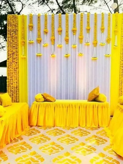 A clean and bright Haldi setup with a white curtain backdrop and yellow marigold hangings. The patterned floor mat adds another layer of design to the space.