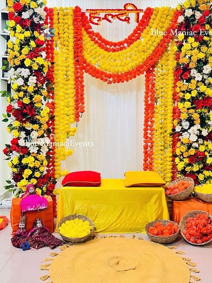A beautiful Haldi setup with a frame of red, yellow, and white flowers and marigold garlands. The traditional Rajasthani puppets add a touch of folk art to the decor.