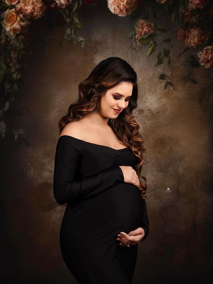 A classic and moody portrait against a painterly backdrop with floral elements. The client's simple black off-shoulder gown and serene expression create a timeless and artistic image.