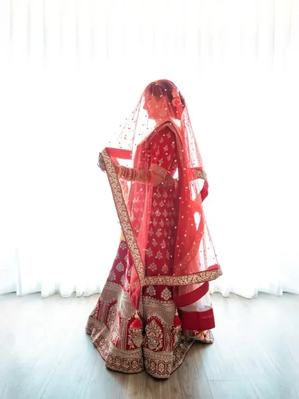 A full-length bridal portrait against a clean, bright background, highlighting the elegance of her red lehenga and veil.