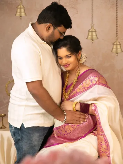 A quiet, intimate portrait of a husband embracing his wife from behind, both focused on the precious life she is carrying, set against a traditional backdrop.