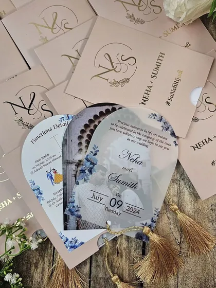 A truly unique fan-shaped invitation that combines modern materials with a playful format. This design features a translucent acrylic card with a delicate blue floral print, attached to a blush pink sleeve with a gold tassel. The sleeve is personalized with the couple's monogram in gold foil.