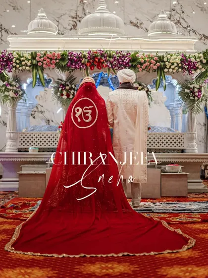 A powerful shot from an Anand Karaj ceremony. The bride's stunning red veil with the 'Ek Onkar' symbol flows behind her as she and the groom stand before the Guru Granth Sahib.