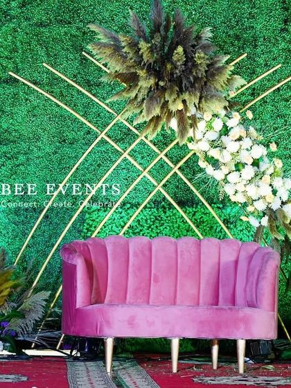 A close-up of our unique reception stage with a green backdrop. The design features a striking diagonal floral arrangement, geometric gold lines, and a chic pink sofa, perfect for a modern and artistic couple.