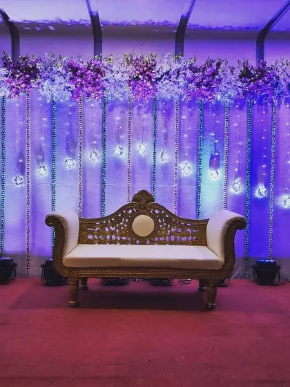 Create a dramatic look with this reception stage rental. It features a classic white sofa set against a backdrop of draped curtains, hanging crystal lights, and vibrant purple uplighting.