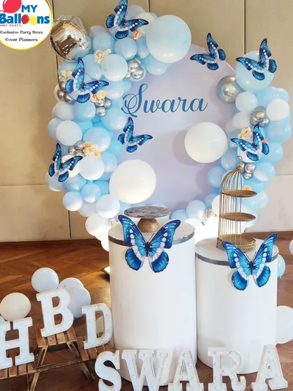 A simple and elegant butterfly-themed setup. The blue and white decor features a circular backdrop with butterfly accents and 'SWARA' marquee letters.
