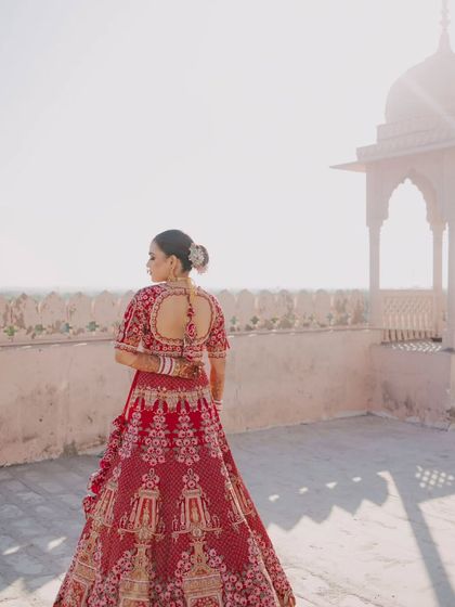 A back profile of the bride, showcasing the beautiful design of her blouse and lehenga against the sunlit palace architecture.