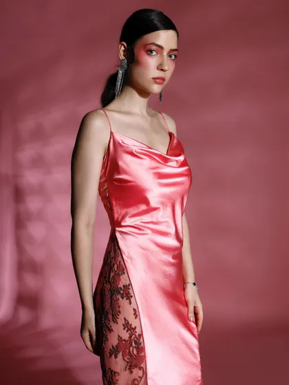 A model in a satin cowl-neck dress with a high slit and lace detailing. The pink background and soft lighting enhance the romantic and feminine quality of the dress.
