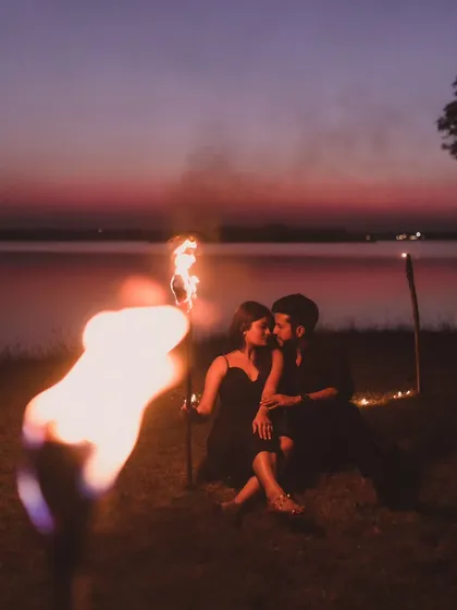 A romantic pre-wedding photo of a couple sitting by a lake at dusk, with the warm glow of a fire torch in the foreground.