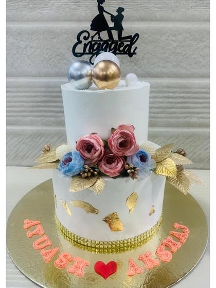 A classic two-tiered engagement cake with a clean white finish, gold leaf accents, and a beautiful arrangement of pink and blue flowers.