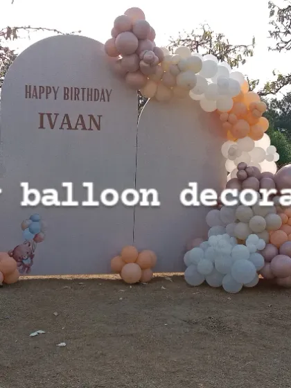 A large outdoor backdrop for a birthday party on a lawn. The setup includes custom arched panels and a balloon garland in a muted, elegant color palette.