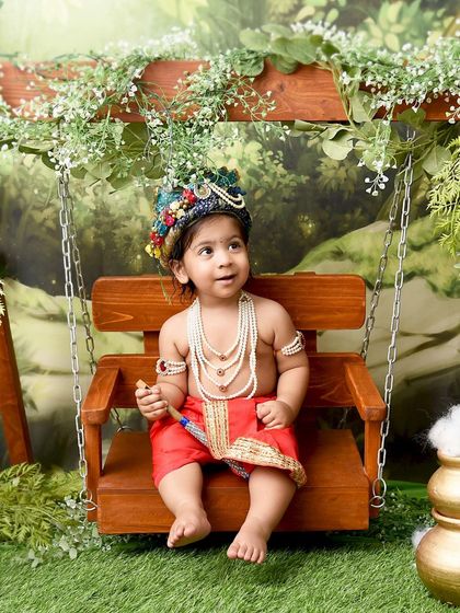 Looking up to the heavens, this little Krishna sits peacefully on a swing. The soft, painterly backdrop and traditional attire create a serene and beautiful image.