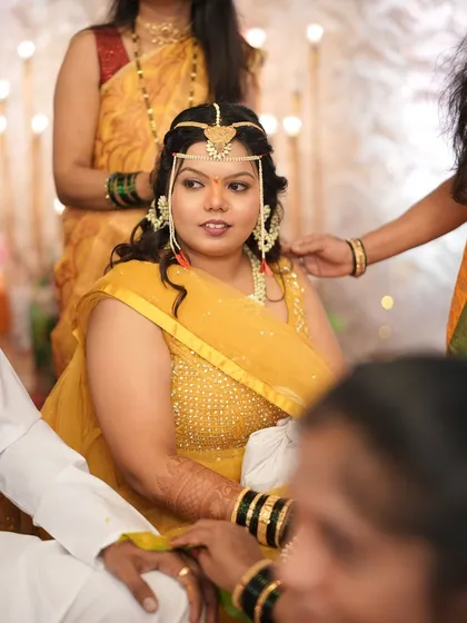 A candid moment from a Haldi ceremony. The bride is wearing a traditional 'mundavalya' made of pearls, complementing her yellow outfit.