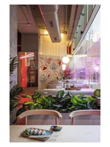 A view from a dining table shows the layers of design, from the lush greenery acting as a space divider to the soft glow of pendant lights and distant neon. This composition creates depth and visual interest throughout the restaurant.