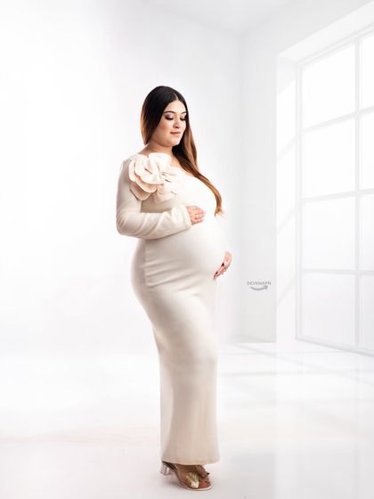 A full-length view of a beautiful, form-fitting cream gown with a floral shoulder detail. The clean, all-white studio setting creates a sophisticated and minimalist fine art maternity portrait.