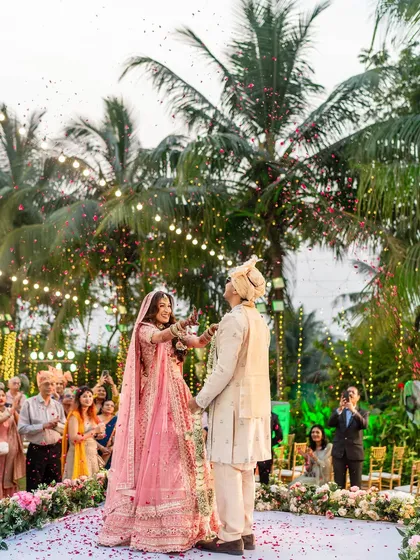 The beginning of the varmala exchange, with the bride playfully trying to garland the groom under a canopy of palm trees and fairy lights.