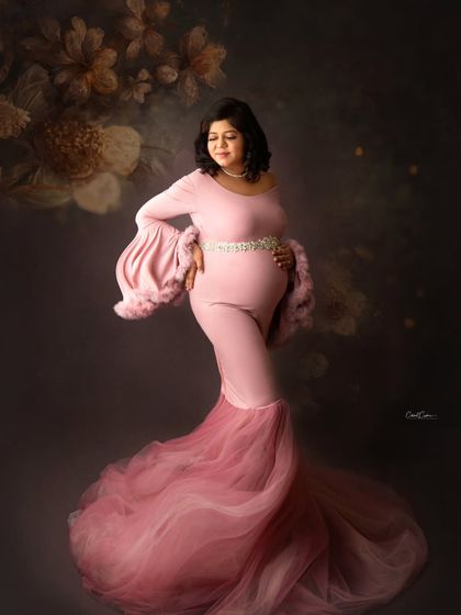 A classic maternity pose showing off the beautiful silhouette of pregnancy. The mom-to-be is in a form-fitting pink gown with flared, feathered sleeves.
