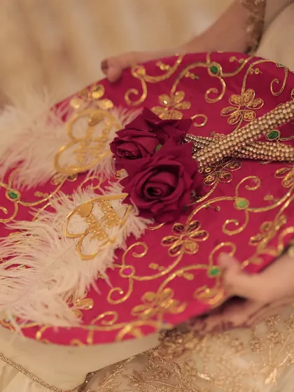 A close-up of a traditional and ornate Nikah ring platter, featuring red fabric, gold embroidery, and delicate white feathers.