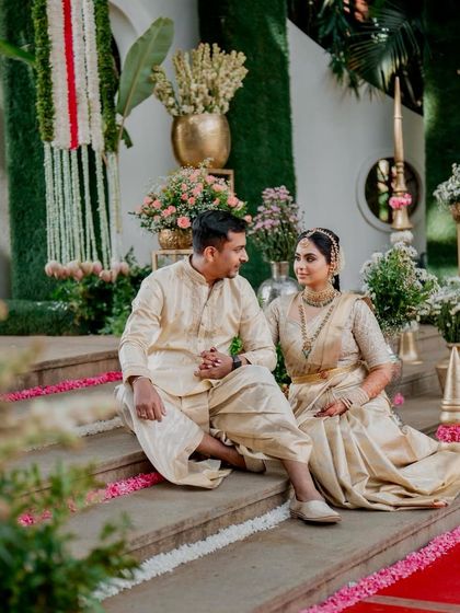 A couple shares a quiet moment on the steps. Their coordinated cream-colored traditional outfits, featuring a silk dhoti-kurta for him and a matching saree for her, were designed for a serene and elegant look.