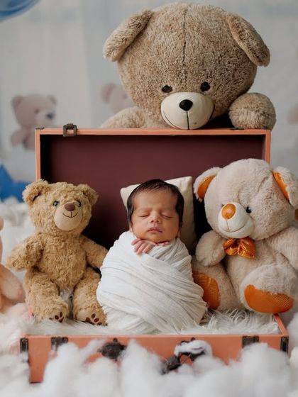 A dreamy and whimsical setup for a newborn. The baby is swaddled and placed in a small suitcase, surrounded by fluffy clouds and a whole family of teddy bears, creating a sweet and imaginative scene.