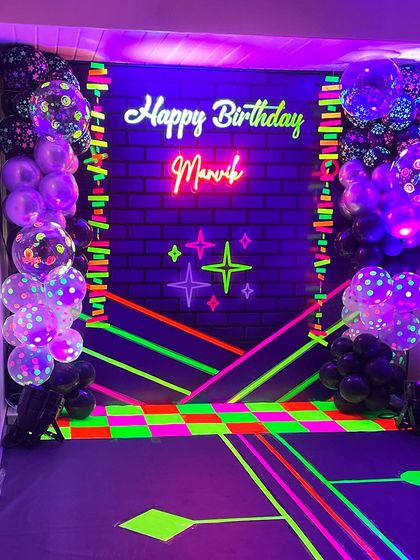 This is how you glow up a party! A complete neon theme setup featuring a custom "Happy Birthday" neon sign against a black brick wall backdrop, surrounded by UV-reactive balloons and floor decals.
