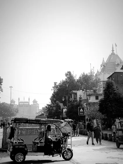 An e-rickshaw passes by with the Red Fort visible in the background, blending old and new transport and landmarks.