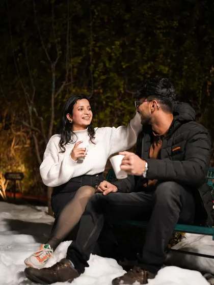 A candid shot of a couple sharing a laugh and a hot drink on a snow-covered bench.