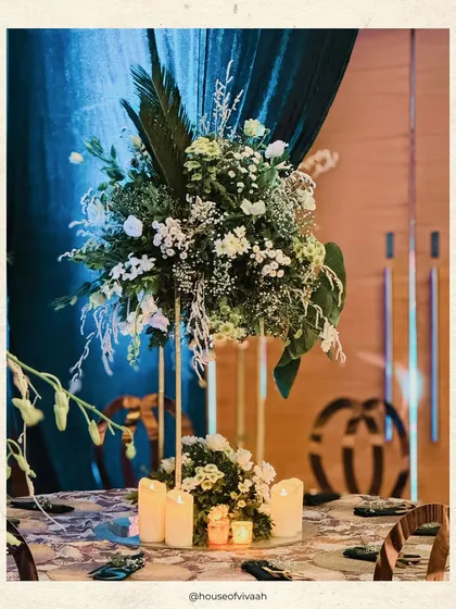A tablescape from a jewel-toned soiree, featuring a tall centerpiece of white florals and lush greens, surrounded by candles, set against a backdrop of emerald drapes.