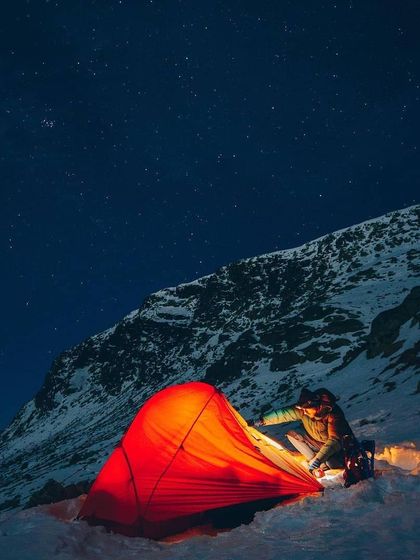 A lone adventurer prepares their glowing tent as night falls on a snowy mountain. The deep blue sky is filled with stars, highlighting the solitude and beauty of winter camping.