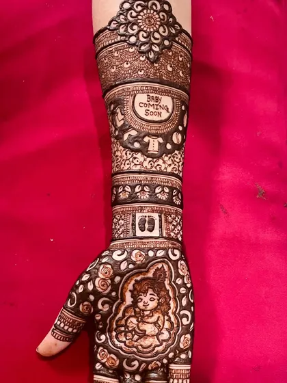A close-up of a baby shower mehandi featuring a portrait of baby Krishna on the palm. The design extends up the arm with motifs like footprints and a 'Baby Coming Soon' message.