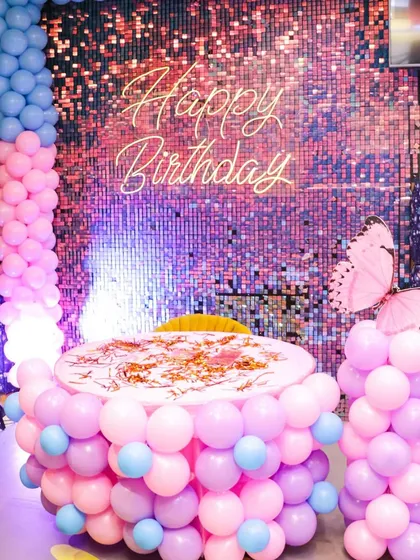 A beautiful butterfly-themed birthday setup. The cake table is wrapped in pastel pink and blue balloons, complemented by a shimmering backdrop and large butterfly accents for a magical feel.