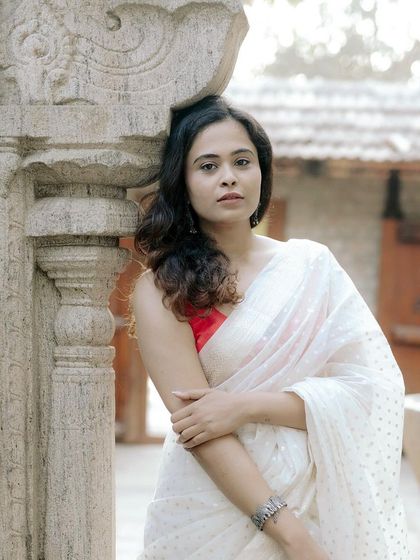 A woman in a simple white saree leans against a carved stone pillar, the soft light creating a timeless and elegant portrait.