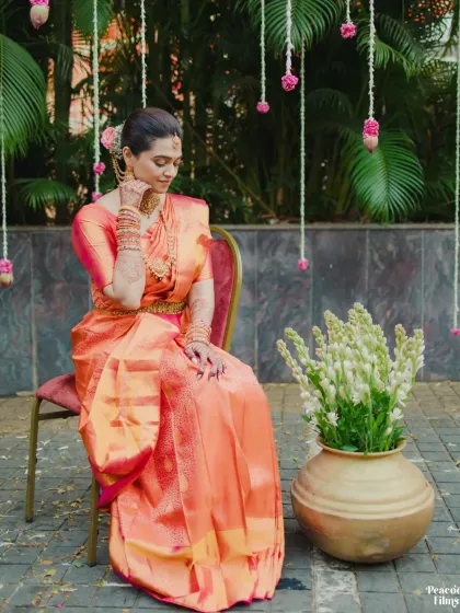 The bride seated in her traditional orange silk saree. This portrait captures the rich colors and textures of her attire and the peaceful setting.