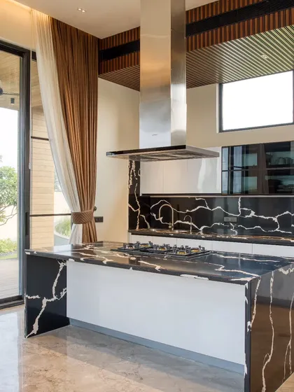 This open-plan kitchen in our Sonipat residence combines high functionality with striking aesthetics. I used a dramatic black marble with white veining for the waterfall island and backsplash, contrasting it with clean white cabinetry and a statement steel chimney for a modern, polished look.