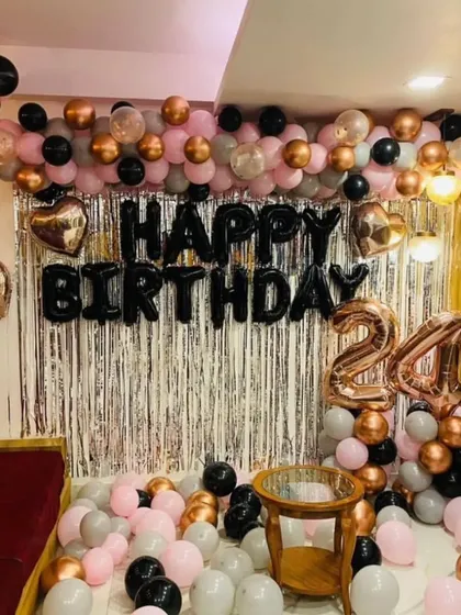 A 24th birthday home decoration with a silver fringe backdrop. The setup includes a balloon garland in pink, black, and rose gold, and foil number balloons.