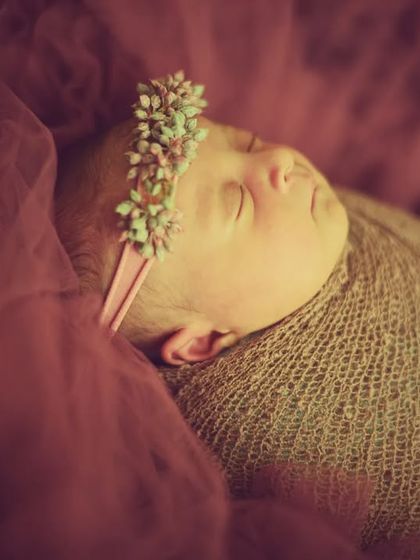 A dreamy, soft-focus portrait. This baby girl wears a floral headband, wrapped in a burlap swaddle against a deep mauve background.