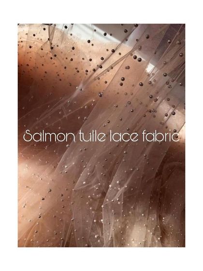 A swatch of delicate salmon tulle lace fabric, dotted with shimmering pearls. This material is chosen for its ethereal quality, perfect for creating couture gowns with a touch of magic.