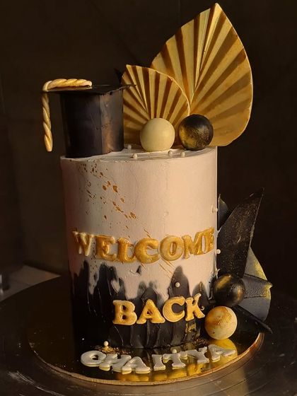 A super pretty graduation theme cake to say 'Welcome Back'. This design features a black and white theme with gold accents, a graduation cap, and custom lettering.